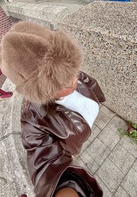 Brązowa skórzana kurtka o błyszczącej powierzchni, w połączeniu z puszystym brązowym kapeluszem. Tło z teksturowanym kamieniem i wzorowanym chodnikiem.