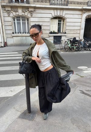 Mujer con gafas de sol, camiseta blanca, chaqueta verde oliva y pantalones anchos negros, de pie en la acera junto a un poste, sosteniendo una pierna levantada detrás de ella.