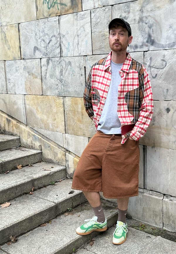 Camisa de franela a cuadros en rojo, marrón y crema; camiseta gris; pantalones cortos marrones; zapatillas verdes y blancas; de pie en unos escalones de piedra contra una pared texturizada.