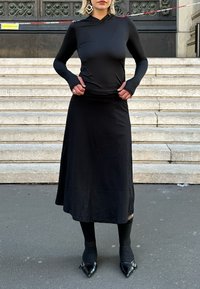Top preto de mangas longas com capuz, design ajustado; saia maxi preta fluida; collants pretos e sapatos de salto agulha com detalhe de fivela.