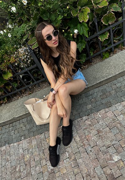 Top negro sin mangas, pantalones cortos de mezclilla, zapatillas de malla negra y un bolso de tote beige. El fondo presenta follaje verde y una barandilla decorativa de metal.