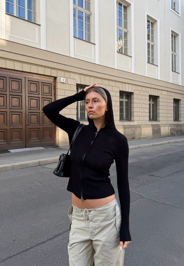 Vrouw die een zwarte hoodie met rits en beige broek draagt, staat op een stedelijke straat met haar hand op haar hoofd, voor een beige gebouw met grote ramen.