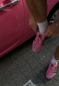 Fehér zoknit viselő személy rózsaszín Adidas sportcipőt húz magára, miközben egy rózsaszín autó mellett áll, szürke, textúrált betonon.
