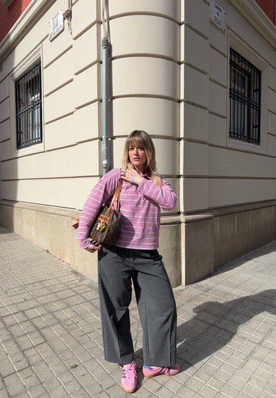 Mujer que lleva una camiseta de rayas rosas, pantalones grises de pierna ancha y zapatillas rosas, de pie en una esquina de la calle iluminada por el sol, sosteniendo un bolso marrón.