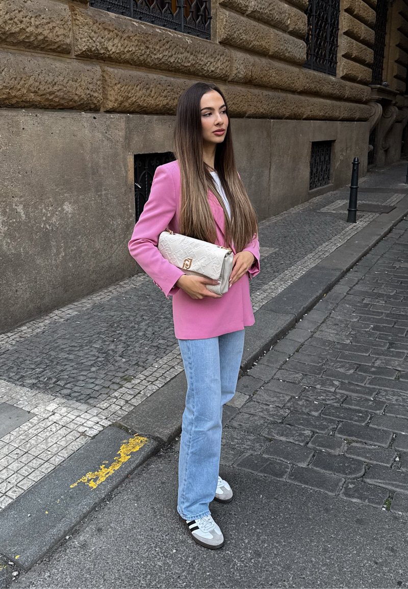 Femme aux longs cheveux portant un blazer rose, un jean bleu clair et des baskets blanches, tenant un sac blanc, debout sur le trottoir d'une rue en ville.