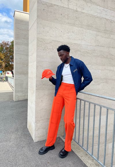 Hombre con pantalones naranja brillante, chaqueta azul marino y zapatos negros se apoya en una barandilla sosteniendo una gorra naranja contra una pared de piedra clara al aire libre.