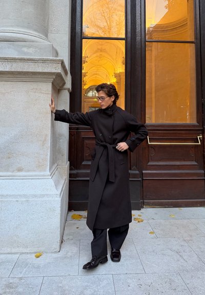 Abrigo trench negro con botones cruzados y cinturón de lazo, combinado con pantalones y zapatos negros, sobre un fondo de pared de piedra.