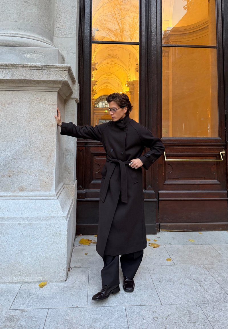 Casaco preto estilo trench coat com botões duplos e cinto de amarrar, combinado com calças e sapatos pretos, colocado contra uma parede de pedra.