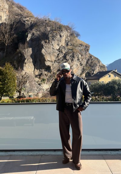 Hombre con gafas de sol, chaqueta de cuero, camisa blanca y gorra de pie en un balcón con una ladera rocosa y casas al fondo bajo un cielo despejado.