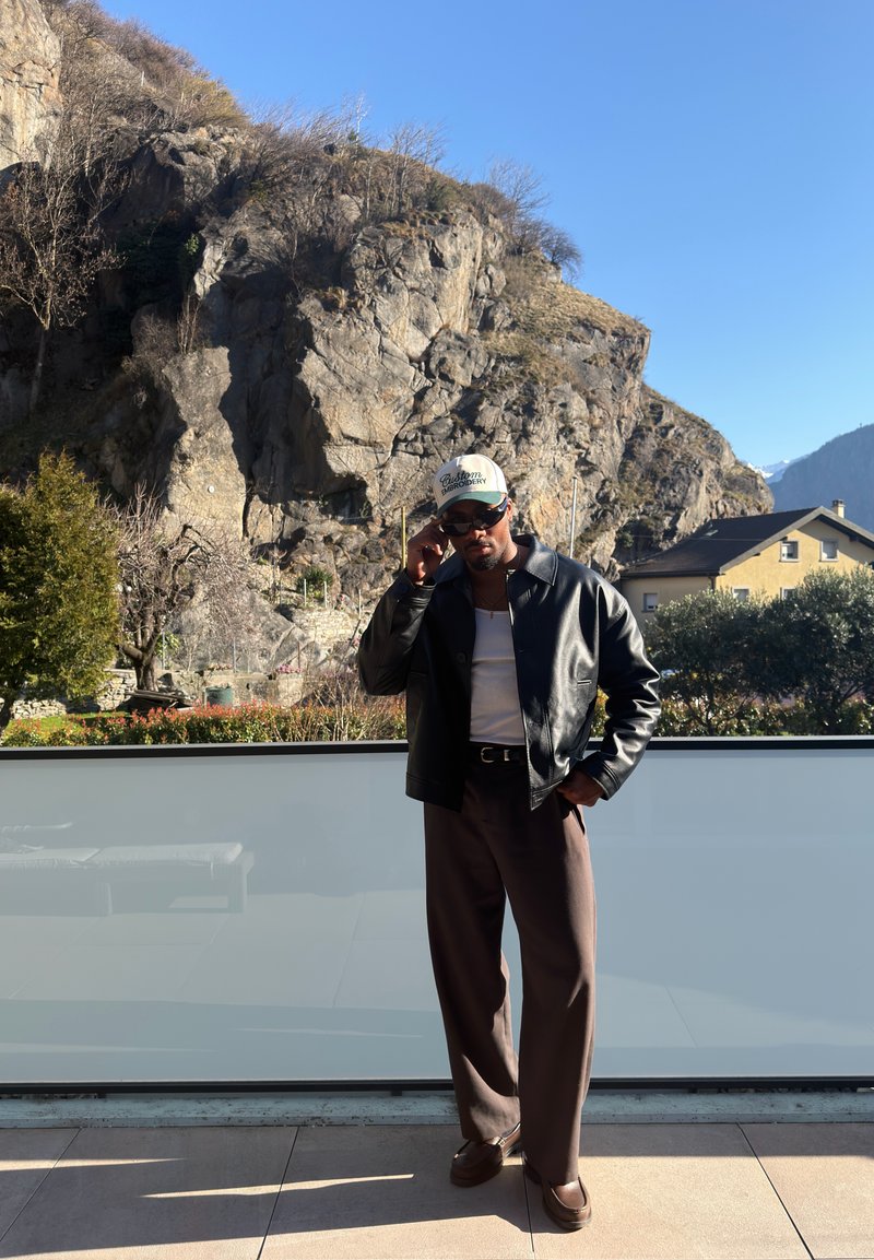 Homme portant des lunettes de soleil, une veste en cuir, une chemise blanche et une casquette, debout sur un balcon avec une colline rocheuse et des maisons en arrière-plan sous un ciel dégagé.