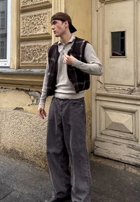 Textured brown plaid waistcoat worn over a light grey long-sleeve shirt, paired with loose grey trousers and a brown cap. Standing against a textured wall.