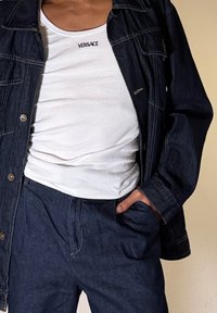 Άτομο που φοράει λευκό τοπ Versace κάτω από σκούρο τζιν μπουφάν και τζιν παντελόνι που ταιριάζει, με το ένα χέρι στην τσέπη.