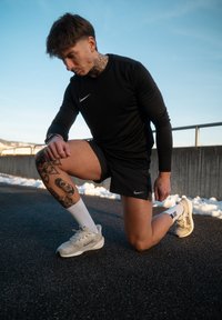 Άνδρας με μαύρη αθλητική ενδυμασία και λευκά αθλητικά παπούτσια γονατίζει στο ένα γόνατο πάνω σε άσφαλτο με χιόνι και κάγκελα στο φόντο.