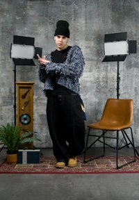 Jeune homme portant un bonnet noir et une veste texturée, posant entre deux projecteurs de studio, à côté d'une chaise marron, une plante et un meuble de musique vintage.