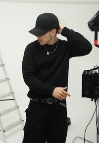 Felpa nera con maniche lunghe, tessuto textured, abbinata a pantaloni cargo neri e una cintura con borchie. È indossato un cappellino nero e gioielli in argento.