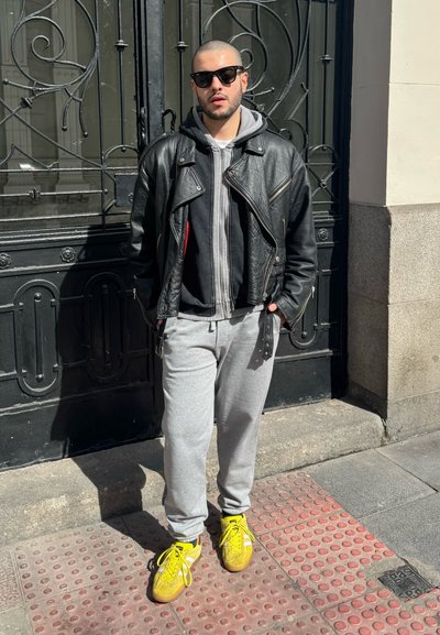 Chaqueta de moto de cuero negro sobre una sudadera gris, pantalones de chándal grises y zapatillas brillantes amarillas. Corte estructurado y atlético con colores en contraste.