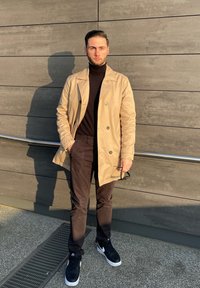 Bež plašč s dvojno zaponko čez črn pulover z visokim ovratnikom, temne hlače in črne športne copate. Plašč ima dvojni gumb in vidne gumbe.