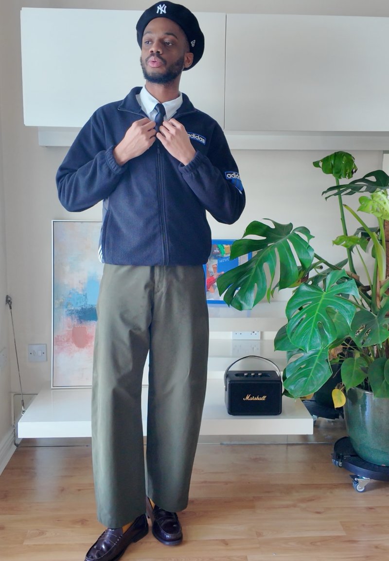 Mann mit schwarzem Hut und Adidas-Jacke, der sein Kragen anpasst, trägt eine Krawatte und olivfarbene Hosen im Wohnzimmer mit einer großen grünen Pflanze und einem Lautsprecher.