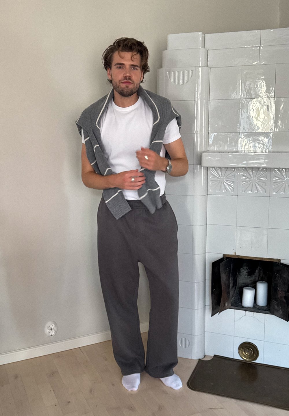 Suéter de lana gris colgado sobre una camiseta blanca, combinado con pantalones grises holgados. Calcetines blancos. Pared de color claro con una chimenea de azulejos.