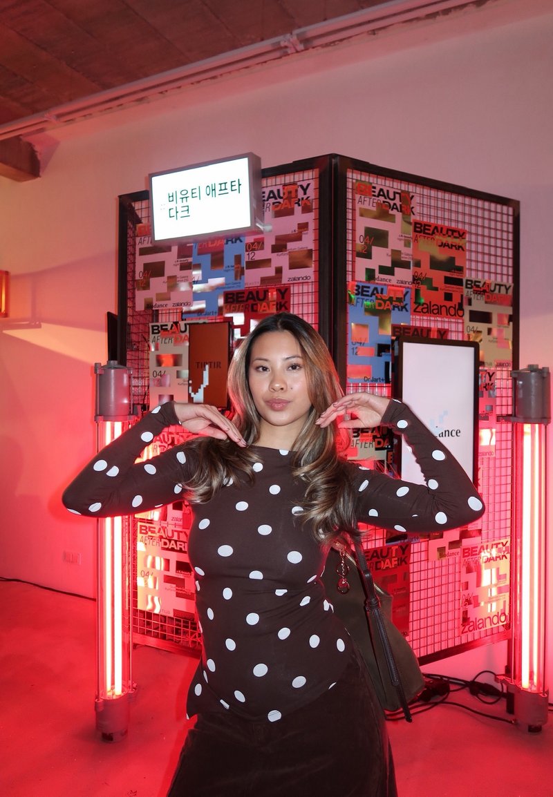 Maglia marrone a maniche lunghe con pois bianchi, abbinata a una gonna scura. Lo sfondo presenta poster colorati e luci al neon.