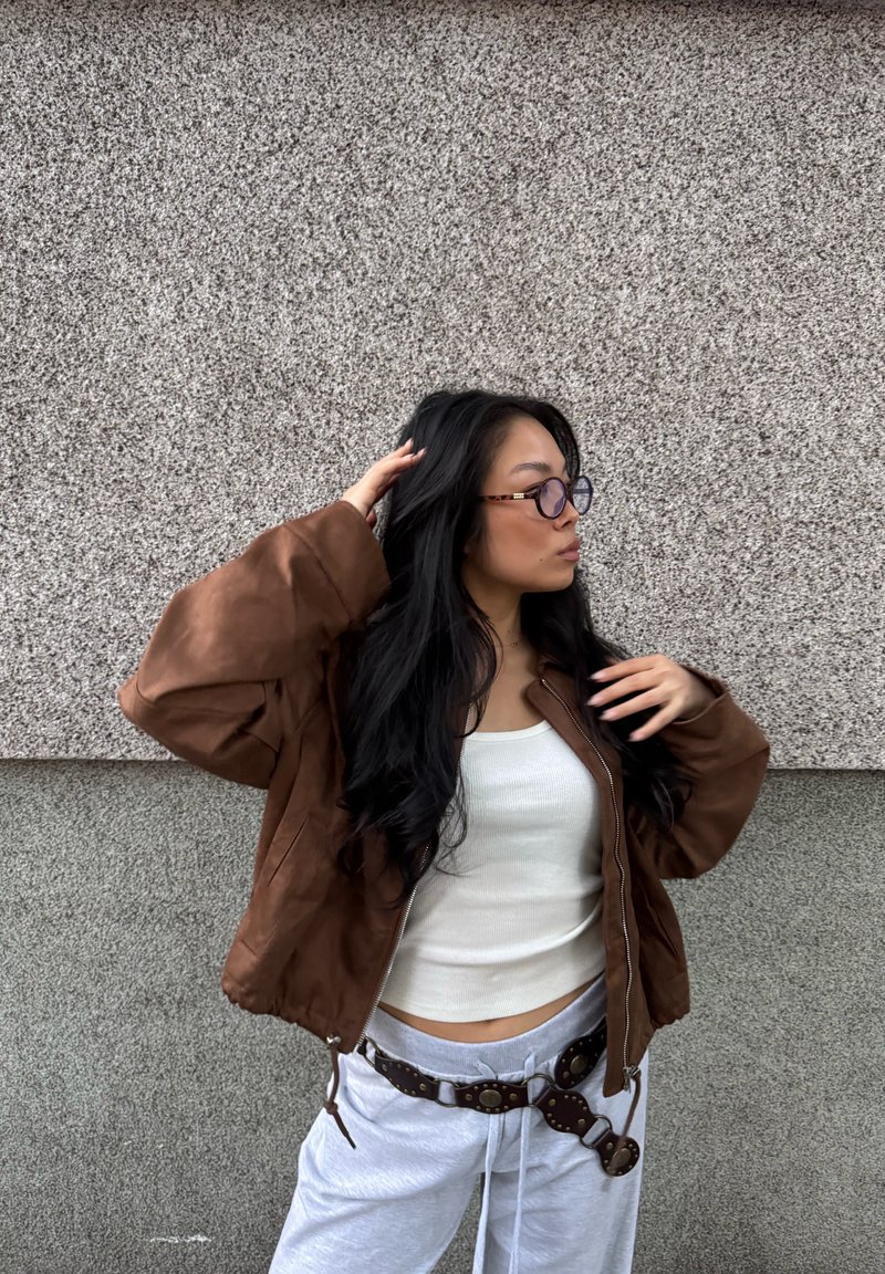 Chaqueta bomber marrón, camiseta sin mangas blanca acanalada, pantalones de chándal grises con un cinturón marrón con tachuelas. Cabello largo y negro, gafas translucidas. Fondo neutral.