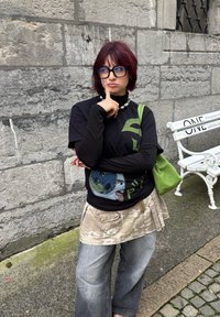 Crna majica s zelenim grafičkim dizajnom, slojevito s bež suknjom s kamuflažnim uzorkom. Široke sive hlače, zelena torba i masivna ogrlica.