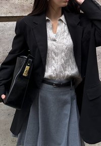 Blazer noir surdimensionné porté sur un pull gris clair à torsades, associé à une jupe plissée grise et un sac à main structuré noir avec des détails dorés.