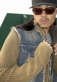 Gilet en denim avec des bords effilochés, associé à des manches côtelées moutarde. Comprend un t-shirt graphique gris en dessous et des lunettes de soleil marron.