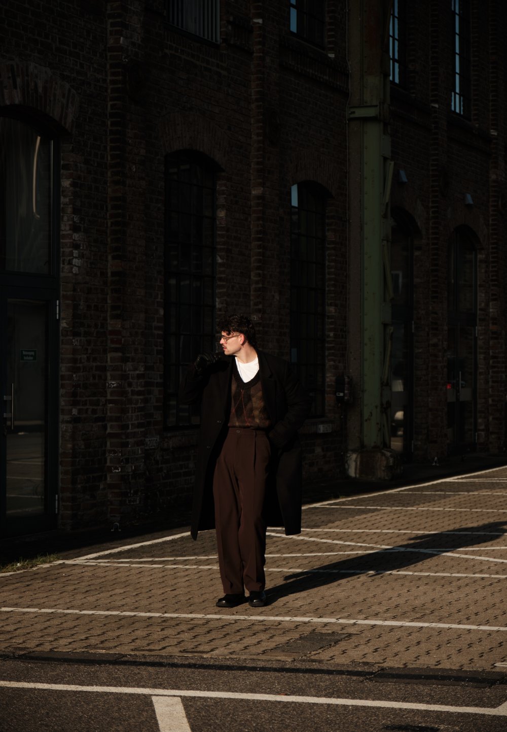 Un homme en manteau foncé, pantalon marron et gants se tient au soleil sur une rue pavée près d'un bâtiment en briques avec de grandes fenêtres.