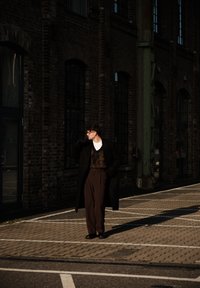 Un homme en manteau foncé, pantalon marron et gants se tient au soleil sur une rue pavée près d'un bâtiment en briques avec de grandes fenêtres.