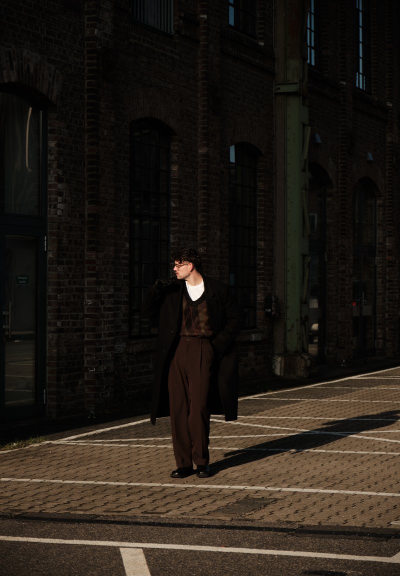 Uomo con cappotto scuro, pantaloni marroni e guanti, in piedi al sole su una strada lastricata vicino a un edificio in mattoni con grandi finestre.