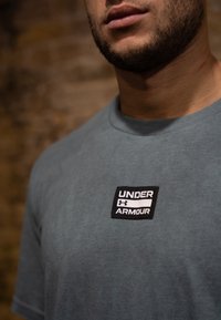 Γκρί βαμβακερό μπλουζάκι με στρογγυλή λαιμόκοψη, που διαθέτει ένα λογότυπο "Under Armour" σε μαύρο και λευκό χρώμα στο αριστερό μέρος του στήθους.