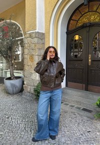 Geacă bomber maro din piele, blugi largi albaștri, ochelari de soare galbeni, pantofi negri, stând lângă un zid de piatră cu o plantă în ghiveci, intrarea vizibilă.