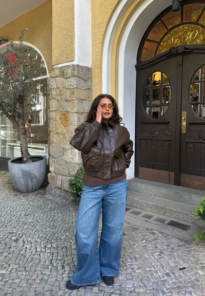 Chaqueta bomber de cuero marrón, vaqueros anchos azules, gafas de sol amarillas, zapatos negros, de pie junto a una pared de piedra con una planta en maceta, entrada visible.