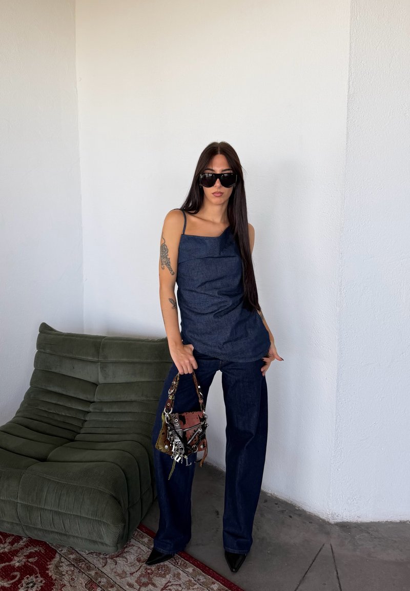 Une femme portant des lunettes de soleil et une tenue en jean foncé pose à côté d’un canapé vert, un sac à main décoré à la main.