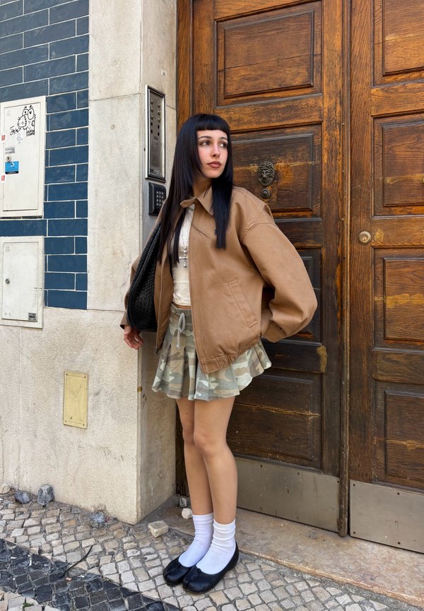 Mlada ženska z dolgimi temnimi lasmi, oblečena v rjavo jakno, maskirno krilo, bele nogavice in črne čevlje, stoji ob lesena vrata na tlakovanem ulici.