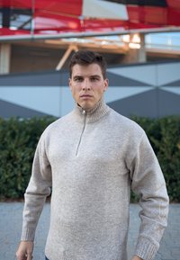 Μπεζ πουλόβερ με φερμουάρ στον λαιμό, φτιαγμένο από πλεκτό ύφασμα, με ριγωτό γιακά και μακριά μανίκια, φορεμένο από ένα άτομο που στέκει μπροστά από γεωμετρικό φόντο.