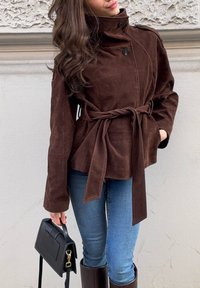 Femme portant une veste marron ceinturée, un jean bleu et des bottes marron, tenant un sac à main noir devant un mur texturé.