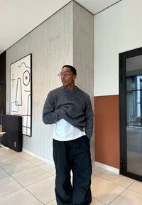 Jovem usando óculos, suéter cinza, camisa branca e calças pretas, está em pé dentro de casa perto de um canto com arte de parede abstrata e uma porta de vidro.