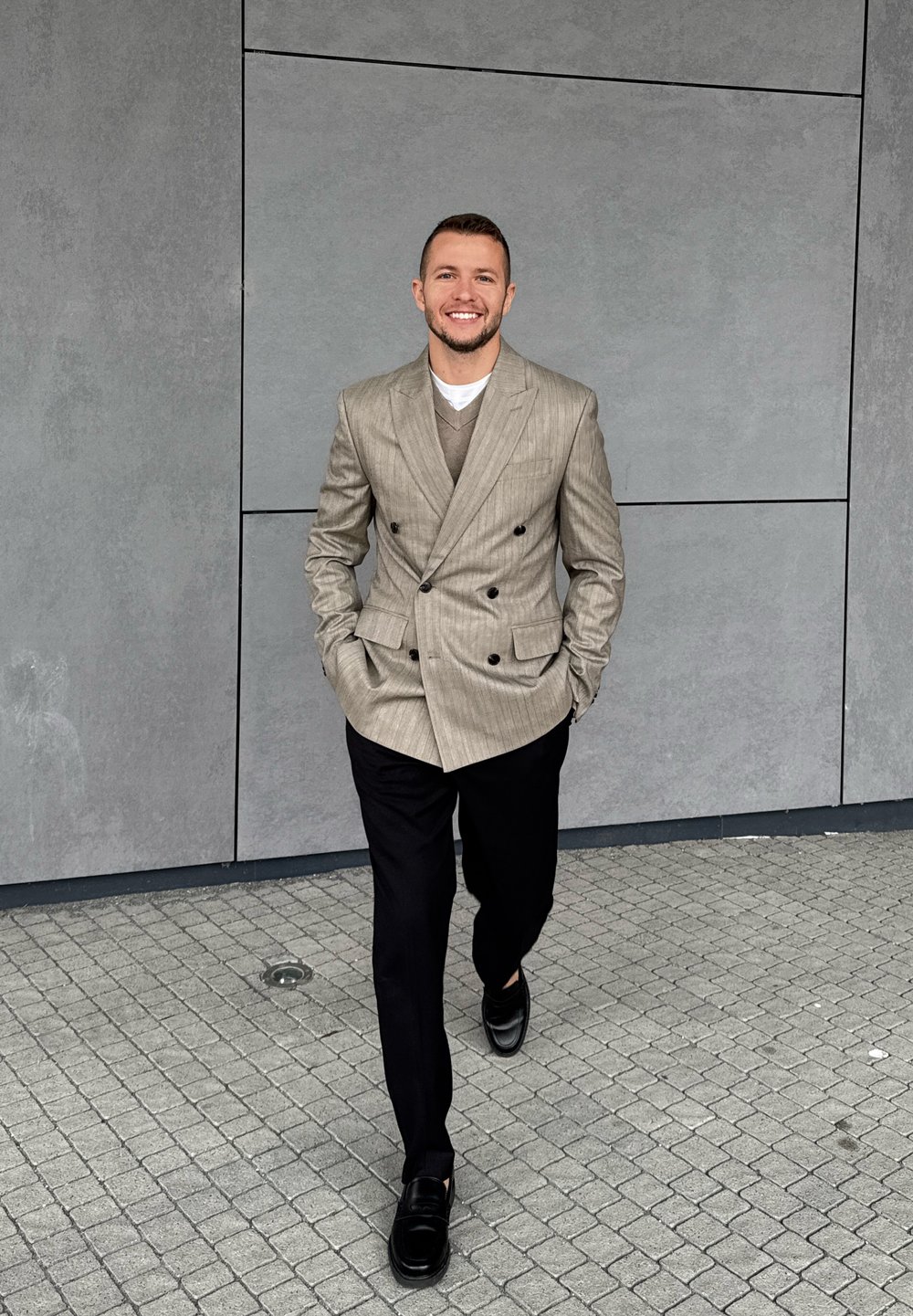 Lichtgrijs dubbel uitgevoerd blazer met verticale strepen, een beige trui eronder, zwarte broeken en glanzende zwarte loafers op een keienoppervlak.