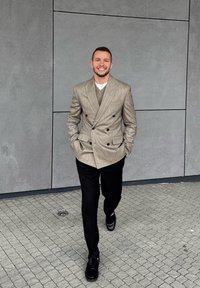 Lichtgrijs dubbel uitgevoerd blazer met verticale strepen, een beige trui eronder, zwarte broeken en glanzende zwarte loafers op een keienoppervlak.