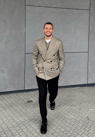 Blazer gris claro de doble botonadura con rayas verticales, suéter beige debajo, pantalones negros y mocasines negros brillantes sobre una superficie de adoquines.