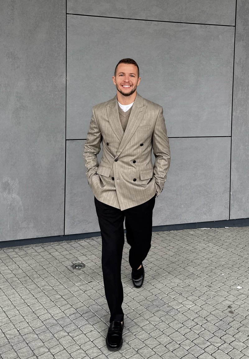 Blazer cinza claro de botão duplo com riscas verticais, sweater bege por baixo, calças pretas e mocassins pretos brilhantes sobre um pavimento de calçada.