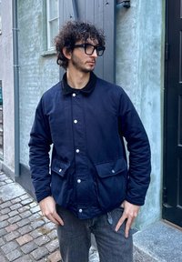 Veste en coton bleu marine avec un col en velours côtelé, des boutons-pression et deux poches avant ; portée sur un jean gris, debout devant un mur texturé.
