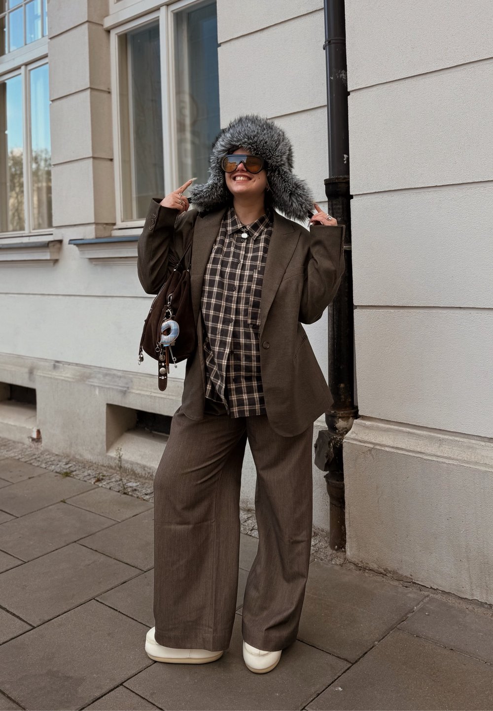 Blazer marron sur mesure porté sur une chemise à carreaux, associé à un pantalon marron à jambes larges et des chaussures compensées blanches. Un chapeau gris doublé de fourrure complète le look.