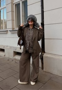 Blazer marron sur mesure porté sur une chemise à carreaux, associé à un pantalon marron à jambes larges et des chaussures compensées blanches. Un chapeau gris doublé de fourrure complète le look.