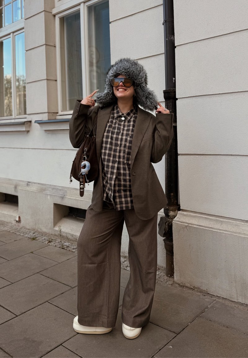 Brauner maßgeschneiderter Blazer über einem karierten Hemd, kombiniert mit weit geschnittenen braunen Hosen und weißen Plateau-Schuhen. Ein grauer, mit Fell gefütterter Hut vervollständigt den Look.