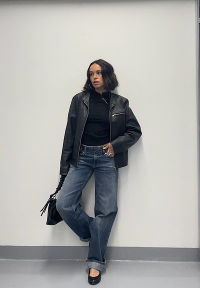 Mujer apoyada contra una pared blanca que lleva chaqueta de cuero negra, top negro, vaqueros azules holgados, zapatos negros, sosteniendo un bolso negro.