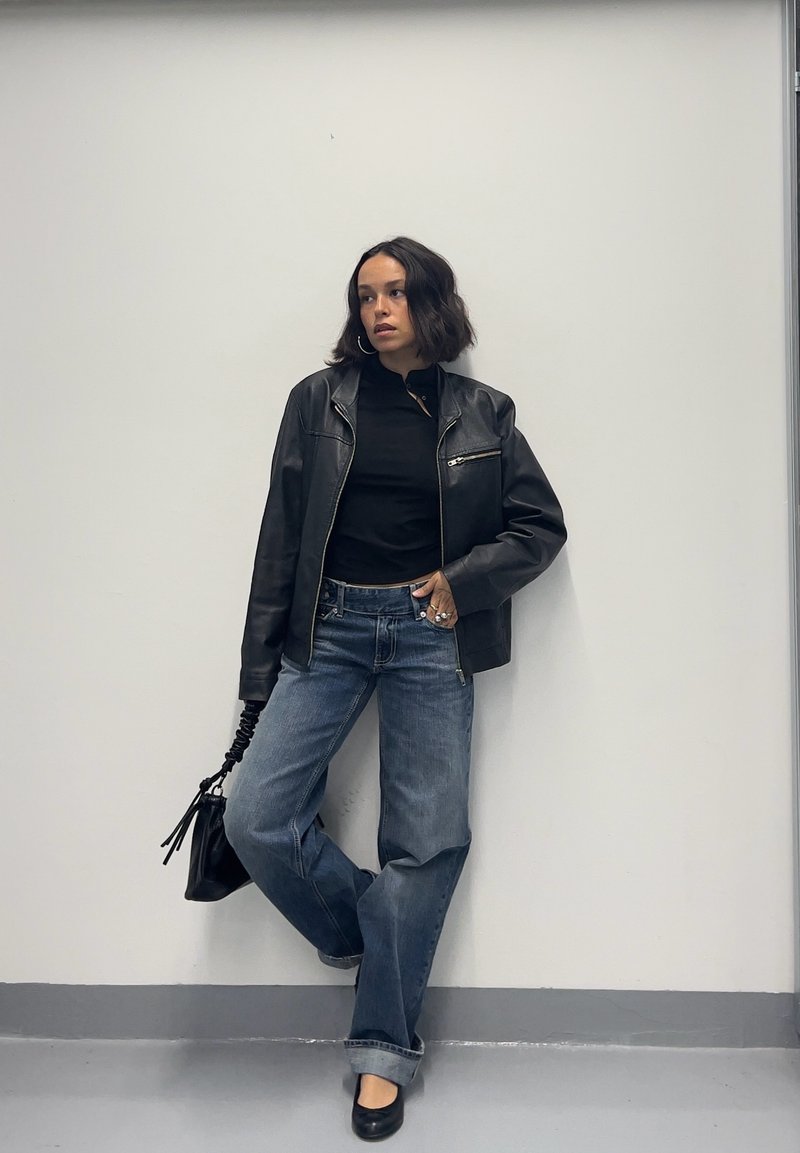 Donna appoggiata a un muro bianco che indossa una giacca di pelle nera, top nero, jeans blu larghi, scarpe nere, che tiene una borsa nera.