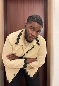 Tânăr cu părul împletit, purtând o jachetă bej cu detalii negre ondulate, cu brațele încrucișate și sprijinindu-se înainte de o ușă din lemn.